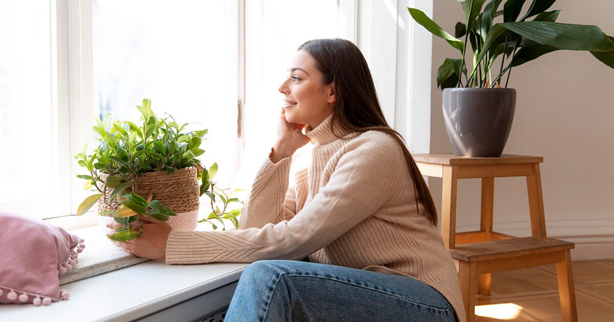 Woman sitting in living room area, surrounded by plants, looking through window.