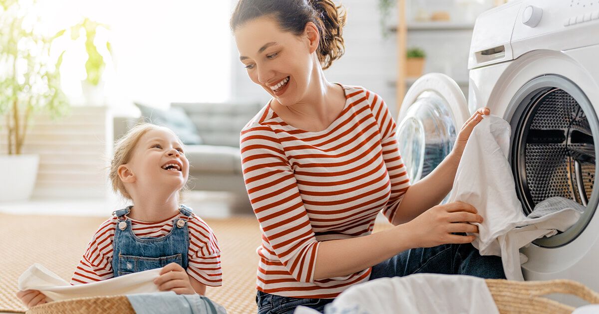 Woman putting clothes in washing machine, smiling at daughter folding laundry.