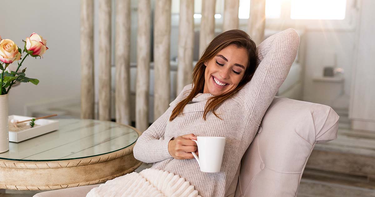 Woman smiling with coffee mug in hand, leaning back on chair in living room.