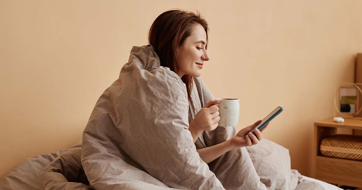 Woman snuggled up in a blanket with a coffee on her phone.